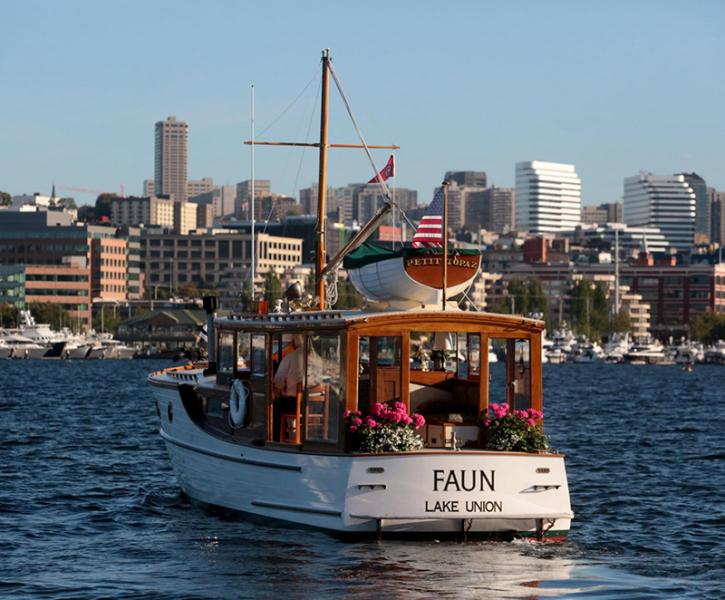 A white boat named “Faun” with “Lake Union” written below the name travels across the water, with pink flowers on the stern and a small dinghy mounted on the cabin roof, set against a backdrop of waterfront buildings and a city skyline.
