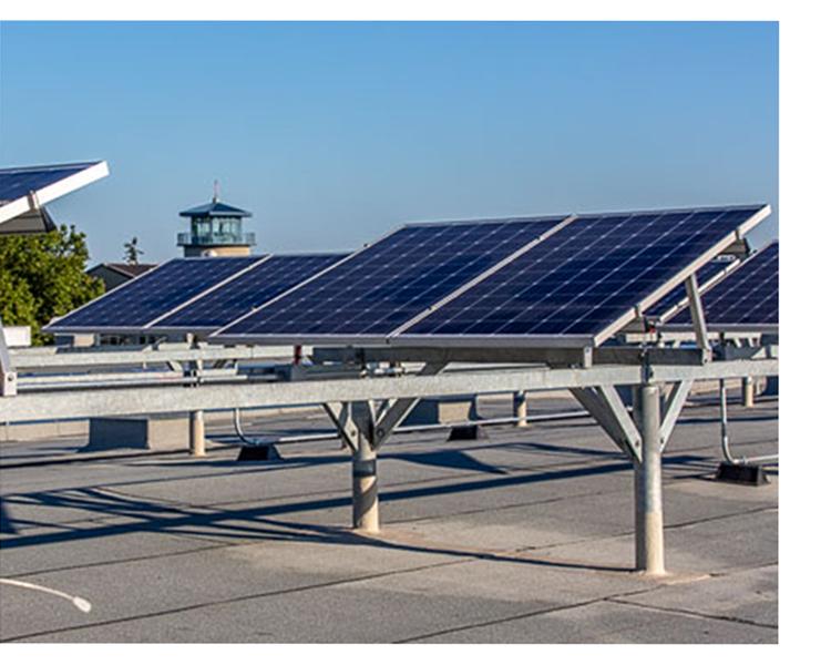 Solar panels mounted on metal frames on a flat rooftop, with a clear blue sky and a small tower structure visible in the background.