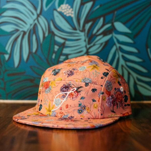 Pink floral-patterned cap displayed on a wooden surface with a blue botanical mural in the background available at Floret