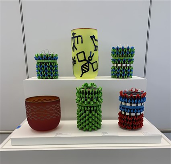 A display of colorful cylindrical glass vessels, including a tall yellow vase with dark blue abstract designs, surrounded by smaller green, blue, red, and brown textured glass pieces arranged on white display platforms.