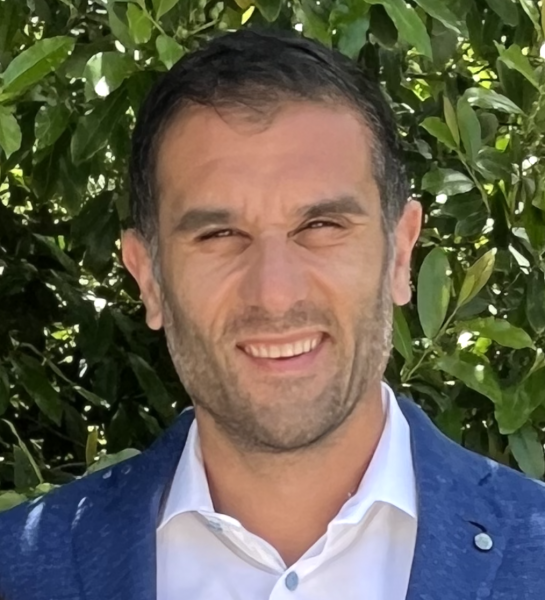 John Salinas wearing a blue jacket and a white shirt is shown outdoors with leafy greenery in the background.