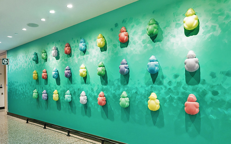 A wall installation titled “wooch isx̱án” in the C Concourse Expansion features multiple rounded, sculptural forms arranged in rows across a bright teal wall. Each form has a smooth, organic shape and is painted in various colors, including green, blue, purple, yellow, red, and white. Overhead lighting casts shadows that highlight the texture and three‑dimensional quality of the pieces. The artwork spans the length of the corridor wall, creating a vibrant and patterned display.