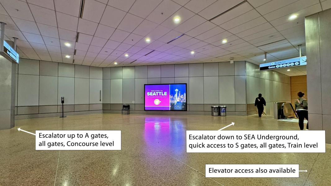 Airport hallway with signs for escalators leading to A gates and SEA Underground, a “Welcome to Seattle” display, and an elevator option nearby.