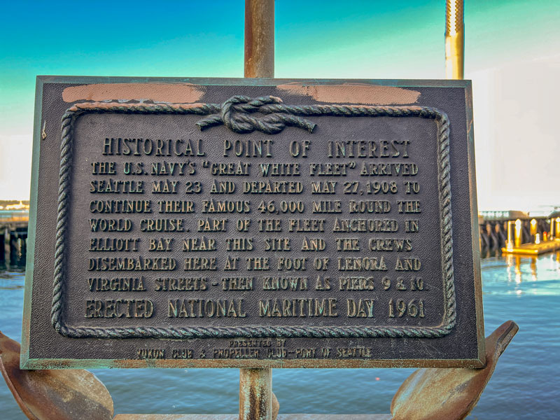 A close-up of a historical plaque describing the visit of the U.S. Navy’s Great White Fleet, mounted beside the water.