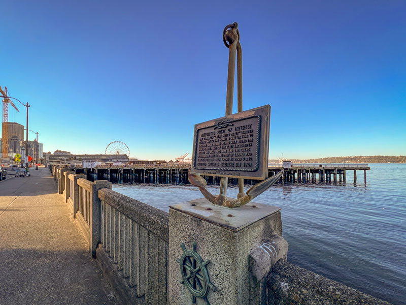 A waterfront walkway with a mounted anchor and historical plaque overlooks a pier and calm water under a clear sky.
