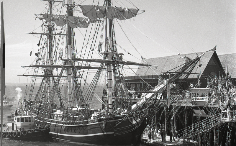 The tall ship HMS Bounty, with multiple masts and detailed rigging, is docked at a wooden pier while a crowd gathers along the structure to watch its arrival.