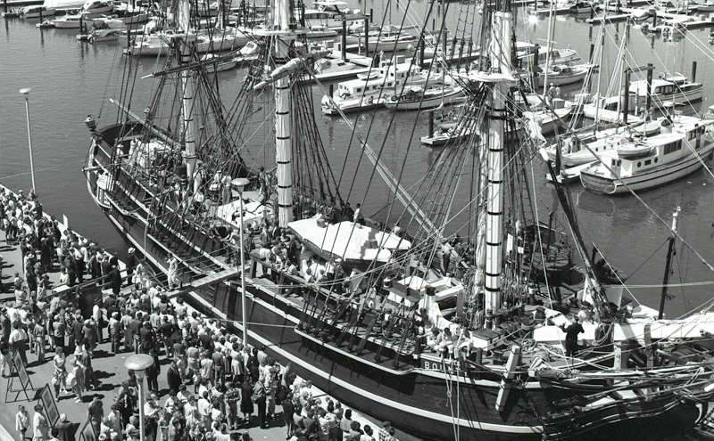 The tall ship HMS Bounty is docked at a marina, with crowds gathered along the waterfront and numerous smaller boats moored in the background.