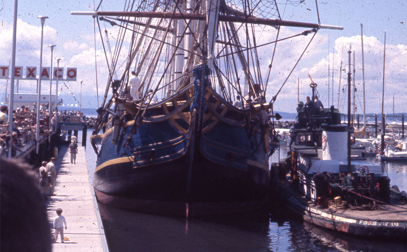 The tall ship HMS Bounty is moored at a dock, with crowds gathered along the pier and a nearby tugboat positioned alongside.