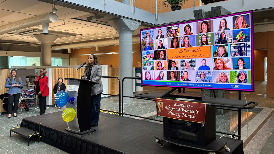 Mohamed speaking at a podium on a small stage in front of a projected collage of female professionals.