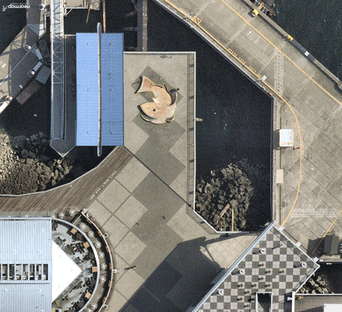 An overhead view of a waterfront pier showing walkways, buildings, a curved plaza area, and water along the edge.