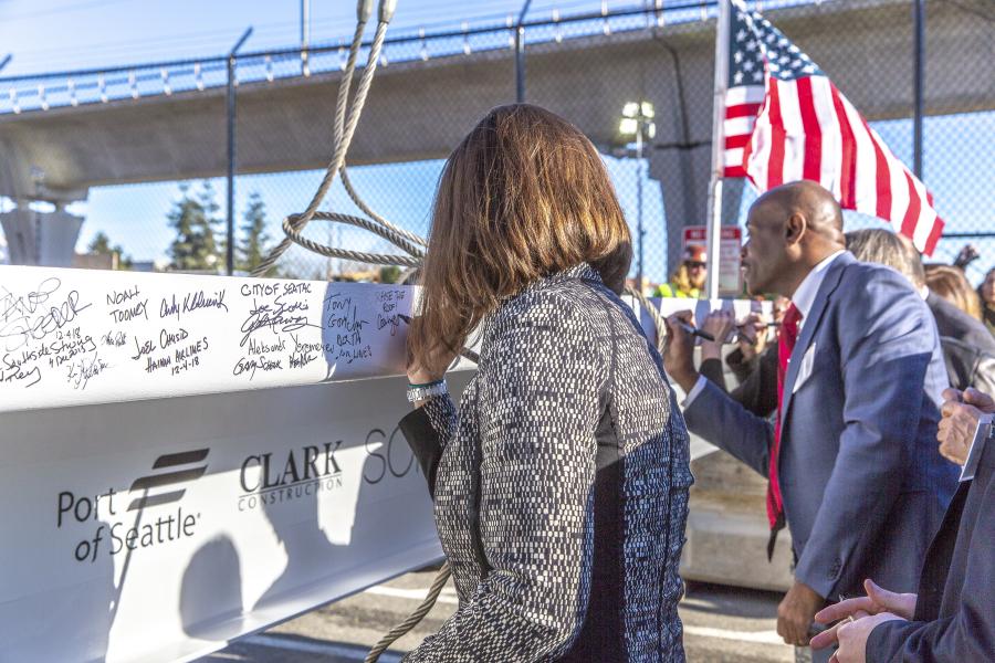 People gather outdoors near a construction beam that is covered in handwritten signatures. A person in the foreground signs the beam while others stand nearby, one holding an American flag. The beam displays logos including ‘Port of Seattle’ and ‘Clark Construction.’ A raised roadway and chain‑link fence appear in the background.
