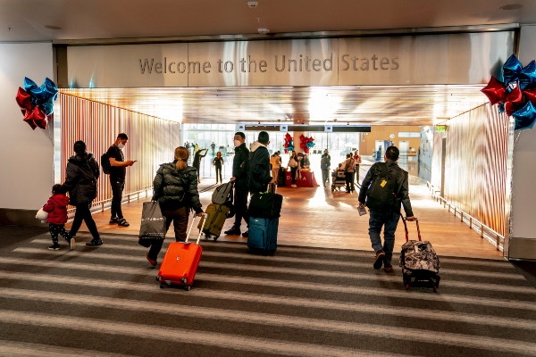 Travelers walk through an airport hallway toward a sign that reads “Welcome to the United States,” pulling luggage and moving toward a staffed area in the distance. The space is decorated with red and blue balloons.