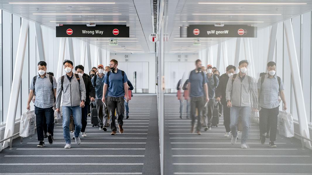 A group of travelers walks down an airport corridor beneath “Wrong Way” signs, with a reflective wall on one side creating a mirror-like duplicate of the scene.