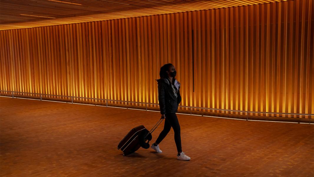 A traveler walks through a dimly lit corridor with warm amber lighting and vertical wall panels while pulling a rolling suitcase.