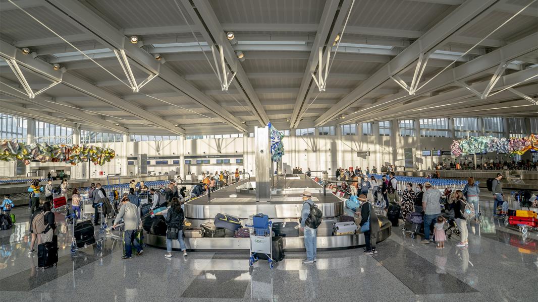 Travelers gather around a large baggage carousel in a bright, spacious airport hall, collecting luggage under high ceilings and large windows.