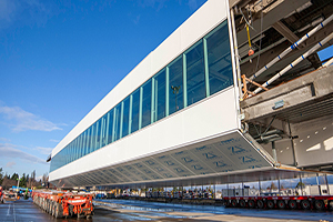 A large prefabricated airport walkway structure being transported across a construction site on heavy-duty wheeled platforms.