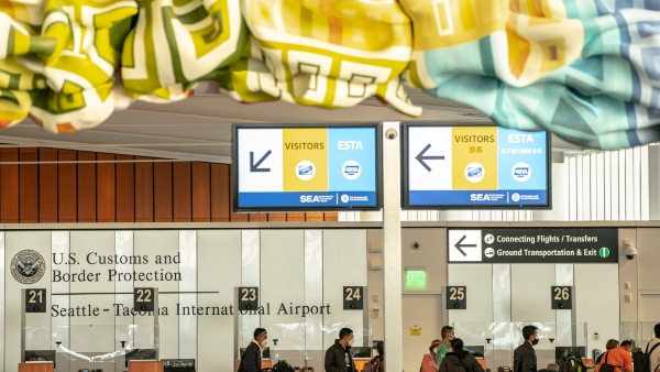 An airport arrivals area shows directional signs for Visitors and ESTA travelers above the U.S. Customs and Border Protection booths at Seattle–Tacoma International Airport, with multiple numbered inspection stations and ceiling art visible.