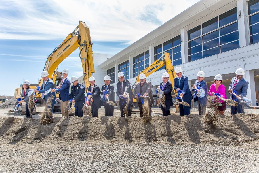 Group of people wearing hard hats stand in a row outside a large modern building, each holding a shovel and lifting dirt in a ceremonial groundbreaking. Two large yellow excavators are positioned behind the group, and the building features tall windows along its exterior. The sky is clear with light clouds overhead.