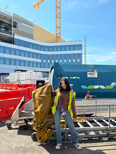 Isabella Newcomer wearing a reflective safety vest stands outdoors beside airport ground equipment and a large blue waste container, with a construction site and crane visible in the background