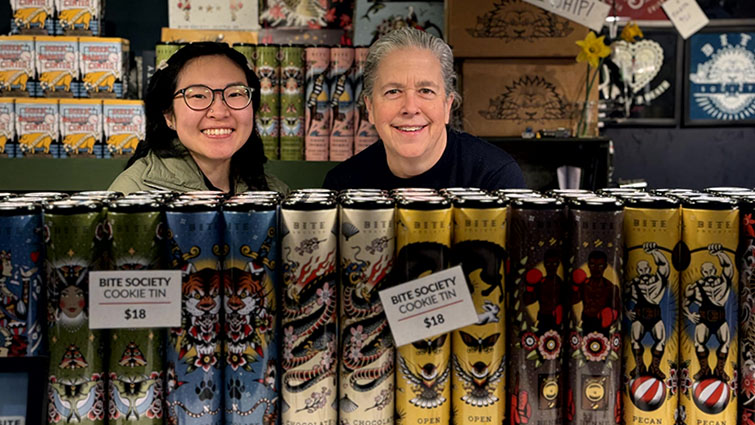 Lendy and staff member pose behind their cookie tins at Pike Place Market.