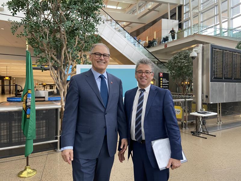 Governor Jay Inslee and&nbsp;Port of Seattle Commissioner Fred Felleman  standing together in an airport terminal near a Washington State flag and signage.