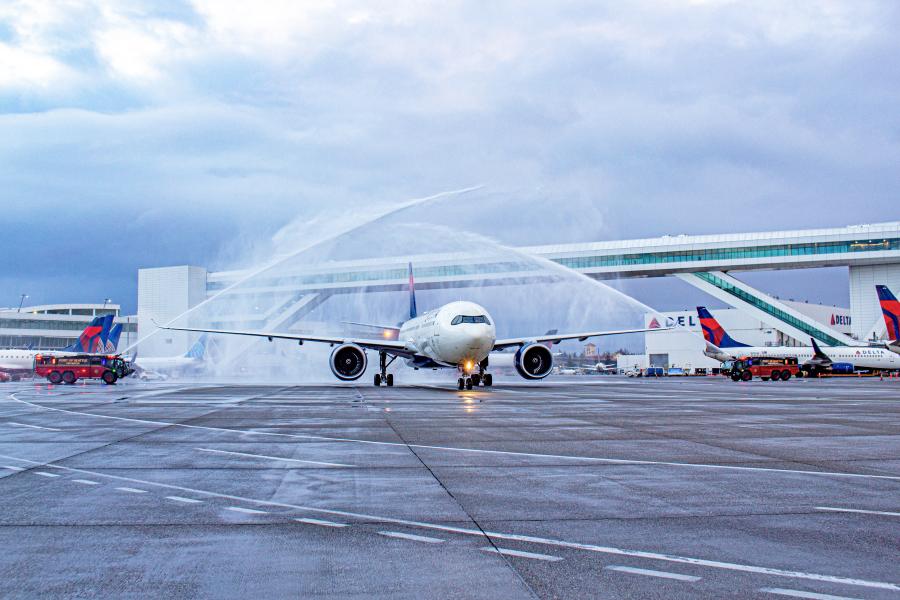 An airplane on an airport tarmac receives a ceremonial water salute from two fire trucks spraying large arcs of water over the aircraft. Several other airplanes are parked nearby, and an elevated passenger walkway stretches across the background. The sky is overcast with gray clouds.