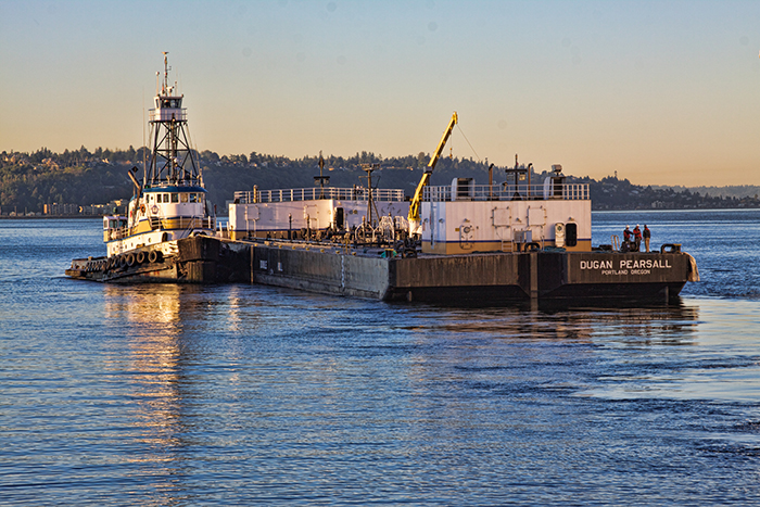 A tugboat guiding a large barge across calm water at sunset, with distant shoreline and trees in the background.