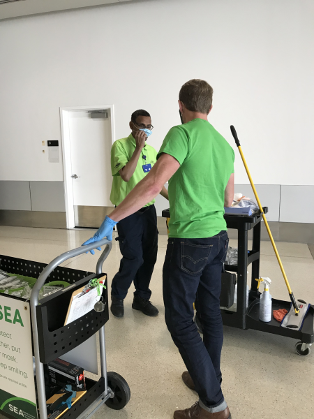 Two people wearing green SEA Airport shirts stand in a hallway, talking near cleaning carts. One person is holding the handle of a supply cart with SEA Airport messaging, while the other is standing beside a janitorial cart with cleaning tools and bottles.