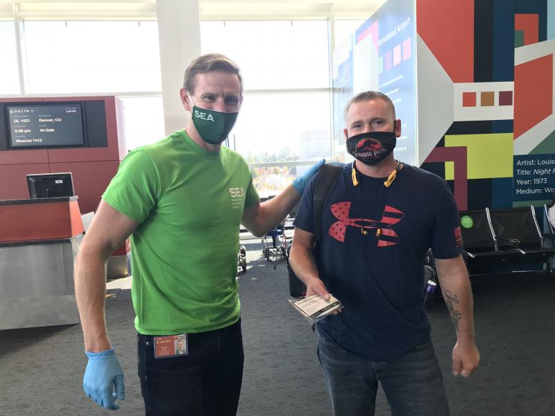 Two people stand in an airport gate area near a seating section and a colorful wall mural. One person is wearing a green SEA Airport shirt and gloves, and the other is holding travel documents. A Delta gate display is visible in the background.