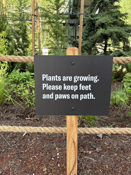 Sign reading “Plants are growing. Please keep feet and paws on path,” mounted on a wooden post with rope fencing and greenery in the background.