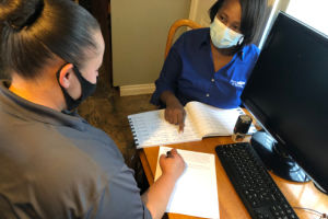 Two people sit at a desk reviewing a notebook, with one person pointing to a page while the other writes on a separate sheet of paper. A computer monitor, keyboard, and mouse are on the desk