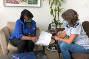 Two people sit in a bright room, reviewing documents at a small table with artwork and a tall plant in the background.