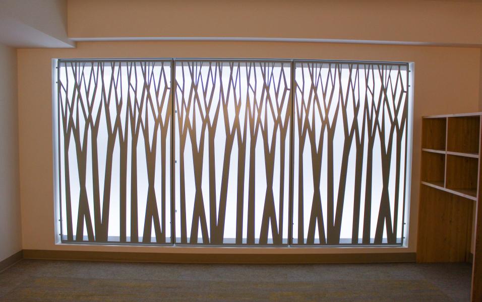A decorative wall panel in the Interfaith Prayer and Meditation Room featuring a pattern of tall, overlapping tree‑like silhouettes, with built‑in wooden shelving visible on the right side of the room.