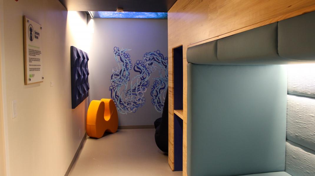 A softly lit sensory room featuring padded seating alcoves, textured wall panels, a blue abstract mural, and a bright orange sculptural chair.
