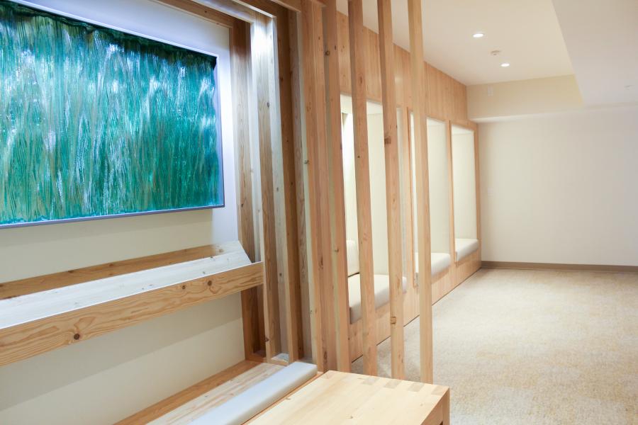 A calm interior space with light wood paneling, built‑in seating alcoves, and a textured green art panel on the wall. The room is softly lit and designed with natural materials and clean lines.