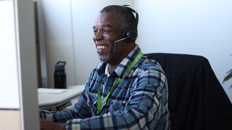 Danan Hoskins wearing a headset and a plaid shirt sits at a desk, looking toward a computer monitor in an office setting.