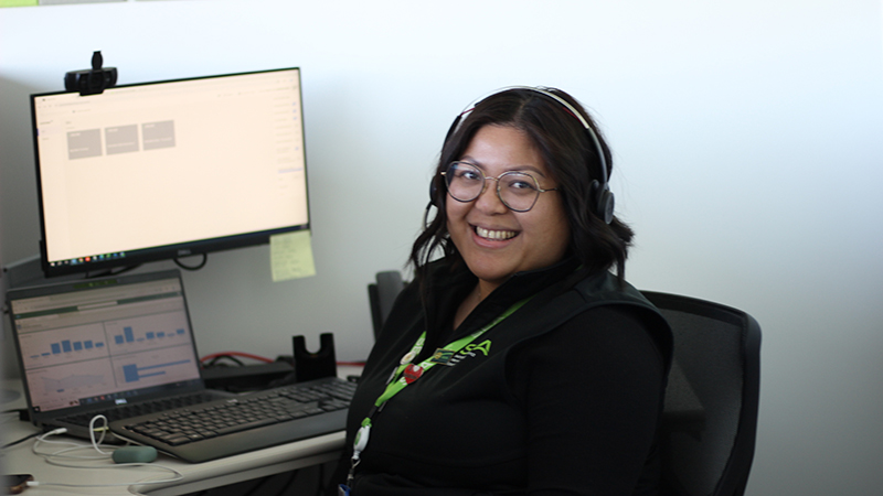 Manuela Taitano wearing a headset and glasses sits at a desk with a laptop and an external monitor displaying charts and interface elements.
