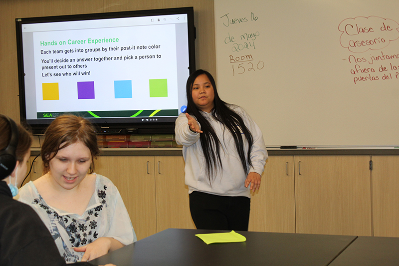A person stands at the front of a classroom extending one arm outward while others sit at a table. A screen behind the standing person displays a slide titled “Hands on Career Experience,” and nearby whiteboards contain handwritten notes.