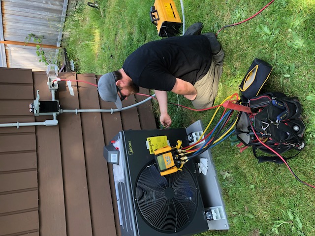 A person kneeling beside an outdoor HVAC unit, using diagnostic tools and connected hoses to perform maintenance.
