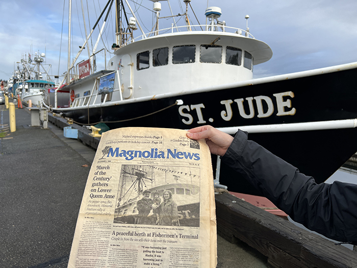 A person holds a Magnolia News newspaper in front of a docked fishing vessel named St. Jude.