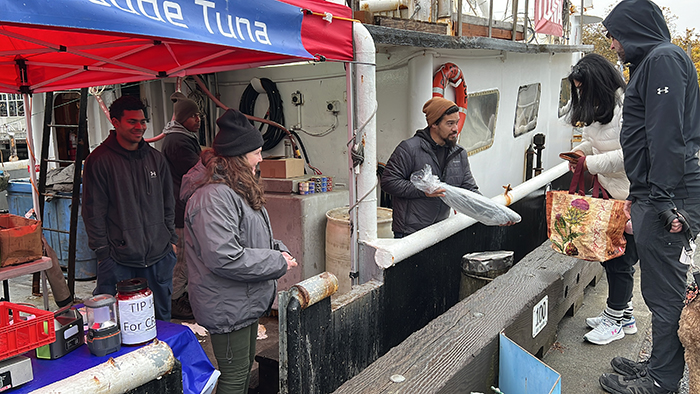 People under a canopy and on a docked vessel exchanging packaged tuna beside a fishing boat.