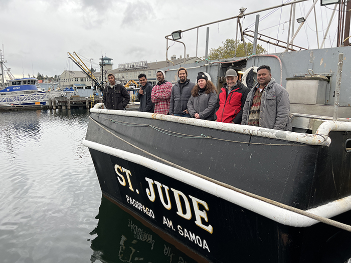 A group of people standing on the deck of the fishing vessel St. Jude, docked beside calm harbor water.