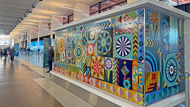 A colorful mosaic inspired by traditional quilting patterns covers a baggage enclosure at SEA Airport, featuring geometric shapes, star designs, and vibrant Pacific Northwest–themed colors.