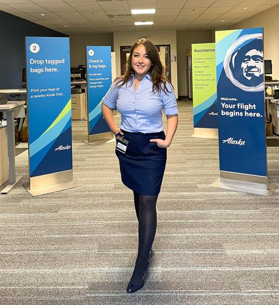 Maddy Knapp in uniform stands in an airport check‑in area between signage displays.