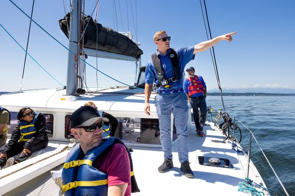 People wearing life jackets on a sailboat, with one individual standing and pointing toward the distance while others sit or stand on the deck.