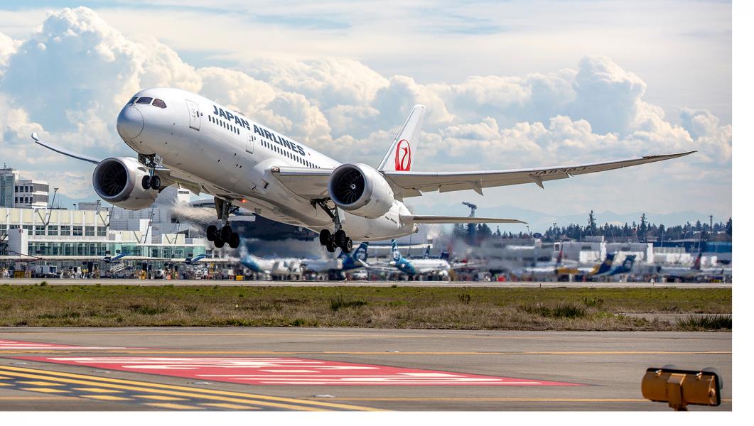 Japan Airlines aircraft lifting off the runway with terminal buildings, ground equipment, and a cloudy sky in the background.