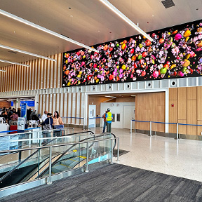 The artwork “Cosmic” by Jennifer Steinkamp is displayed as a large digital screen high on an interior wall, showing a vivid arrangement of colorful floating flowers against a dark background. The scene is set in a spacious indoor area with wood paneling, escalators, and people walking nearby.