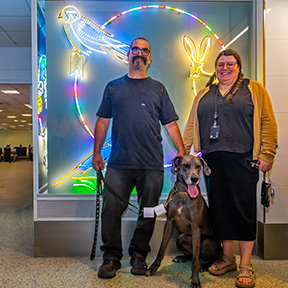 Two people stand indoors beside a large illuminated artwork featuring bright neon-style outlines of birds and plants. A large dog sits in front of them, facing the camera.