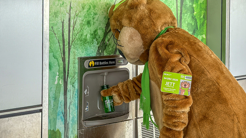 The SEA mascot JETT fills a reusable bottle at a water station with a forest-themed background.