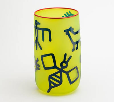 A yellow glass cup with a red rim featuring bold, dark blue abstract line drawings, including geometric shapes and stylized animal-like figures, displayed against a plain white background.